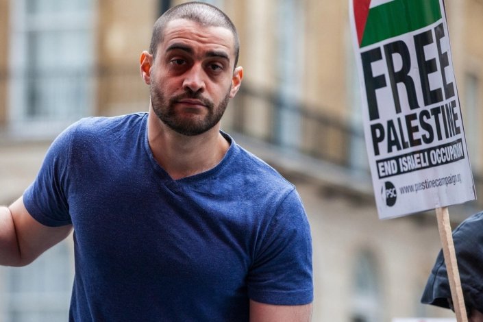 Lowkey ao lado de um cartaz da Palestine Solidarity Campaign