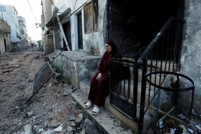 Mulher sentada à entrada de uma casa bastante destruída, numa rua também bastante arrasada, no campo de refugiados de Jenin