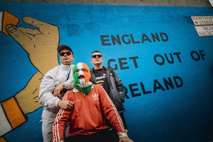 Os Kneecap, um trio de hip-hop da Irlanda do Norte, em frente a um mural onde se lê: Inglaterra, sai da Irlanda!